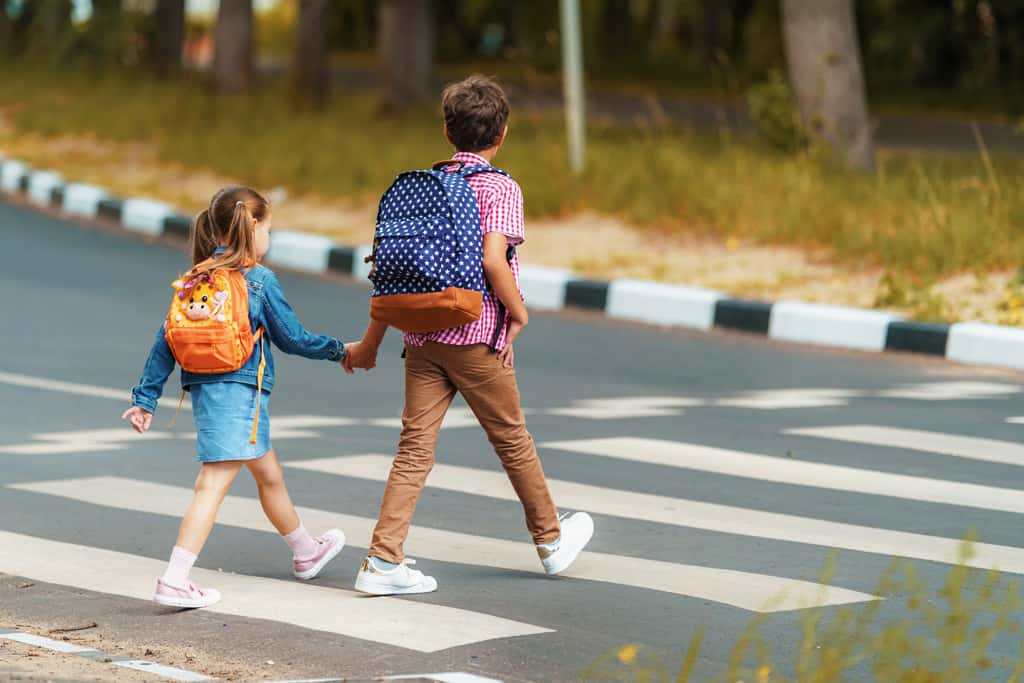 Two children wearing backpacks cross the street at a crosswalk.
