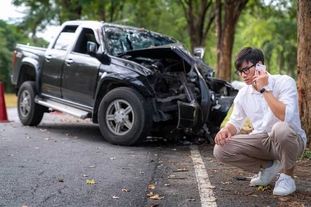 A man crouched on the ground, on a phone, with a black pickup truck visibly crashed from the front.