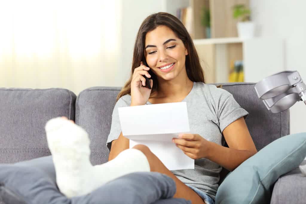 A person sitting on a couch in the living room at home, talking on the phone while reading a letter, with a leg in a cast and crutches resting against the couch.