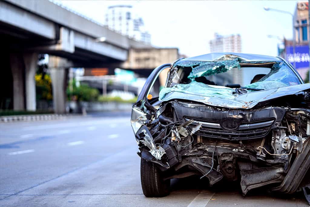 A heavily damaged black car is seen on the side of an urban road, with its front end completely crushed and the windshield shattered.