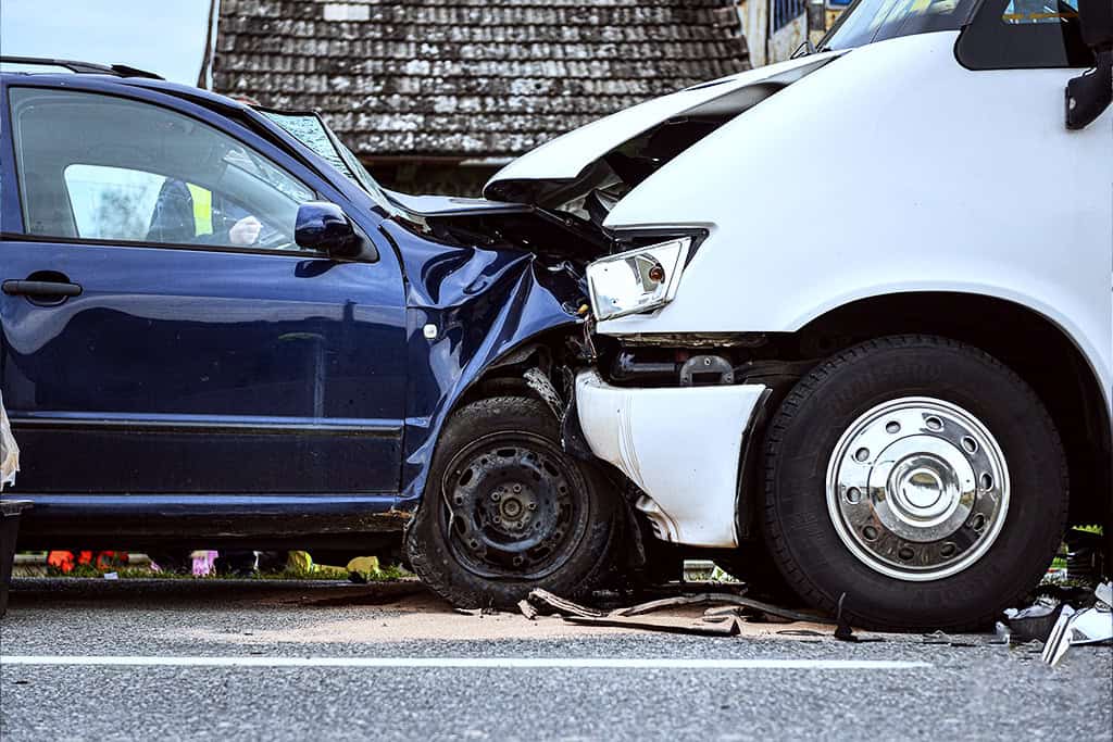 A head-on collision between a dark blue car and a white car. Both vehicles have severe front-end damage, with crumpled hoods and broken headlights.