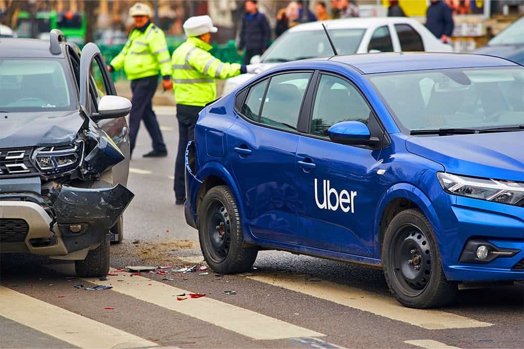 A car accident involving a blue Uber vehicle and a black SUV at an intersection, with visible front-end damage to both cars. Police officers in reflective jackets are managing the scene in the background.