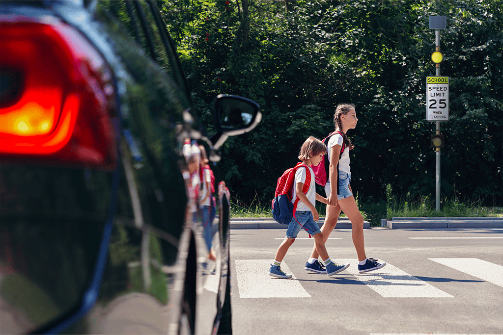 Two children with backpacks crossing a street at a pedestrian crosswalk near a school zone, with a car stopped in the foreground and a school speed limit sign visible in the background.