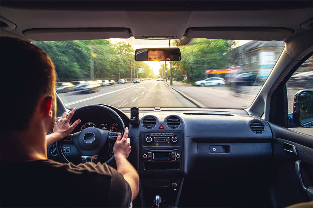 View from the back seat of a car as a man drives at high speed down a city street at sunset, with motion blur showing the surroundings rushing past and a smartphone mounted on the dashboard.