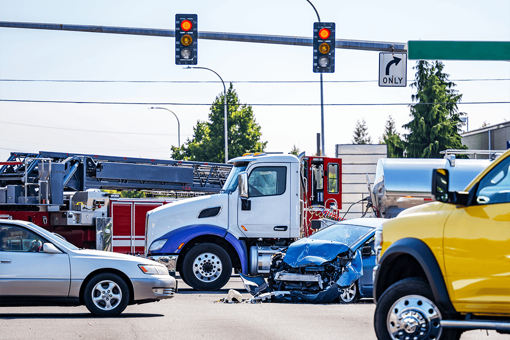 Multiple vehicles are involved in a serious collision at a city intersection, including a blue car with significant front-end damage, a fire truck, a white and blue semi-truck, and several other cars. A traffic light overhead displays a red signal, and emergency responders appear to be on the scene.