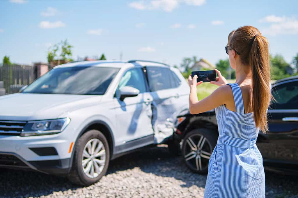 A woman in a blue and white striped dress is taking a photo with her smartphone of two cars involved in a collision.