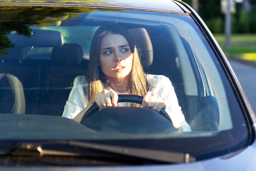 A young woman sitting in the driver's seat of a car, gripping the steering wheel tightly and looking to her left with a worried or anxious expression.