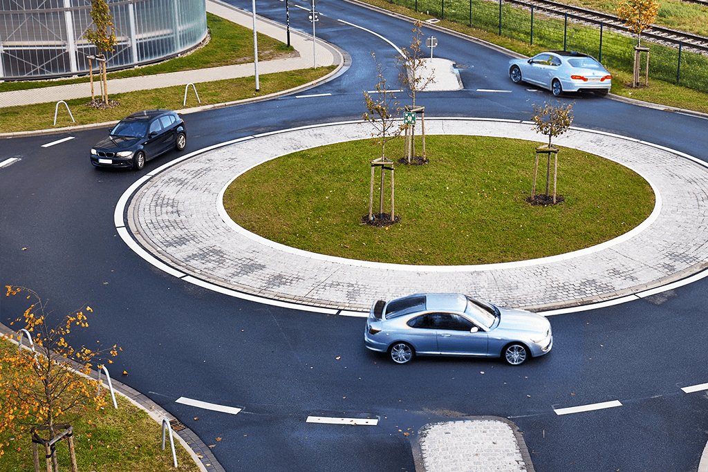 A roundabout with three cars navigating through it on a clear day, surrounded by grass, young trees, and nearby buildings and rail tracks.
