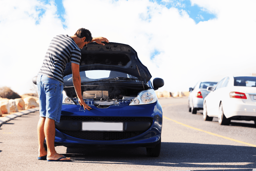 A man in shorts and a striped shirt looking under the hood of a blue car that has broken down on the side of the road, with other vehicles passing by in the background.
