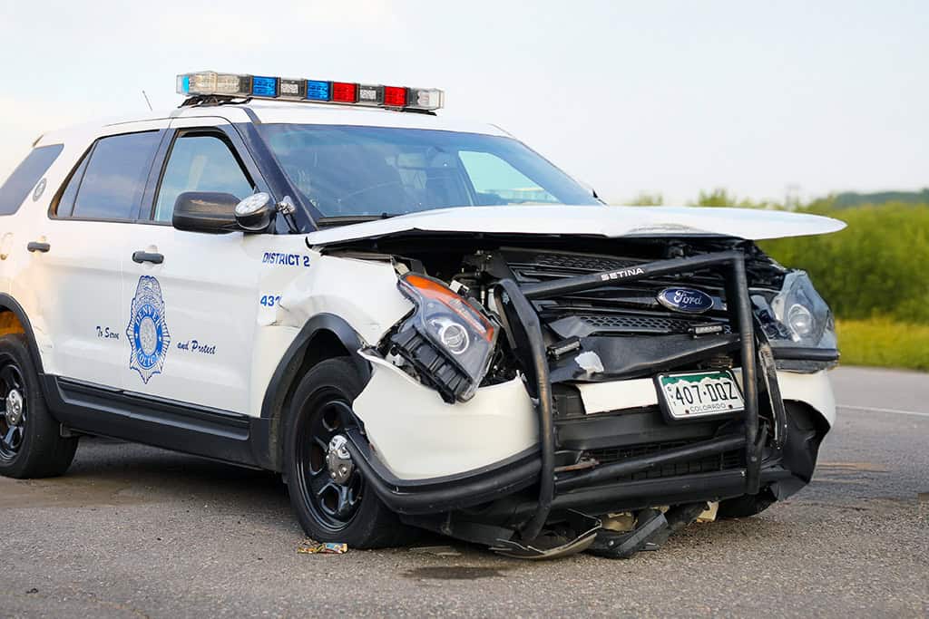 Damaged white police SUV with front-end collision damage, dented hood, and broken headlight, on the road.