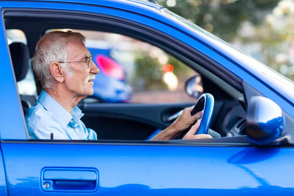 Elderly man wearing glasses driving a blue car, viewed from the side with the window open.