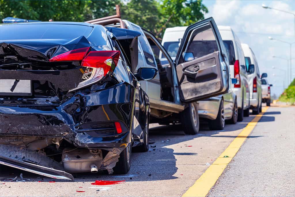Multi-car rear-end collision on a roadway, showing several damaged vehicles lined up with visible dents, broken parts, and an open car door.