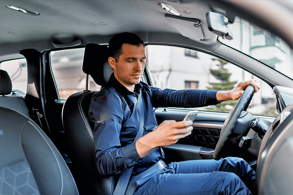 A man sitting in the driver’s seat of a car, wearing a seatbelt and holding the steering wheel with one hand while looking down at a smartphone in his other hand.