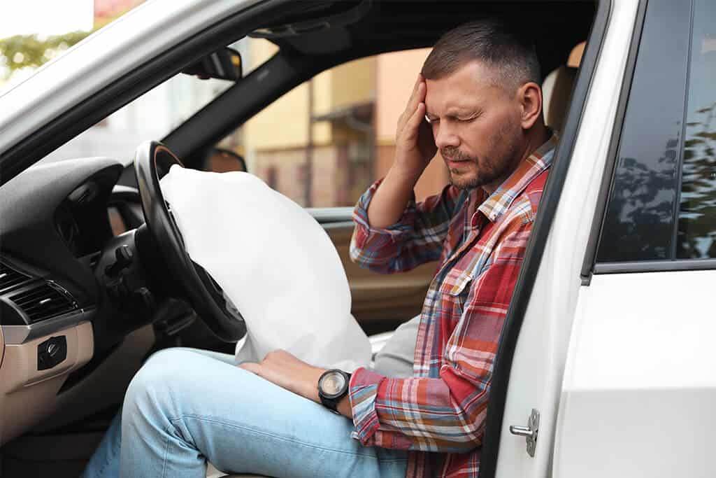 Man with head pain after car accident sitting in vehicle with airbag deployed.