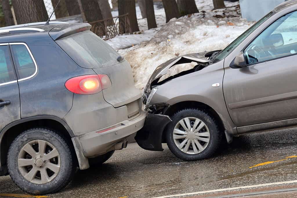 Collision of two cars on the road in winter, with the rear car showing front-end damage and the car ahead displaying brake lights.