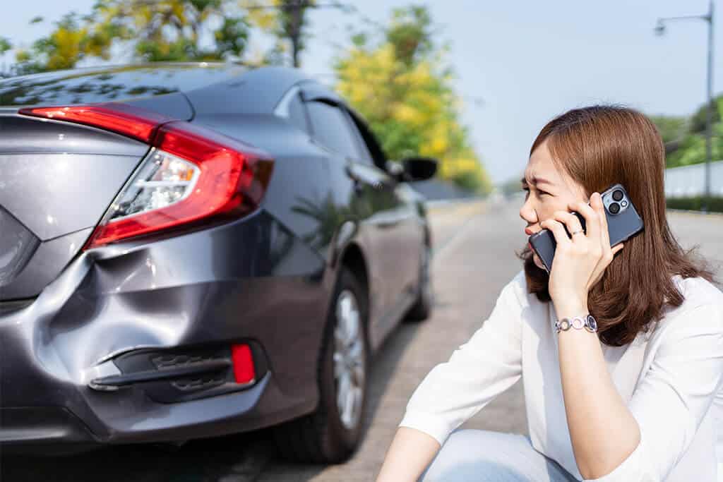 A woman sits beside her vehicle after an accident. She is using her cell phone to call for assistance.
